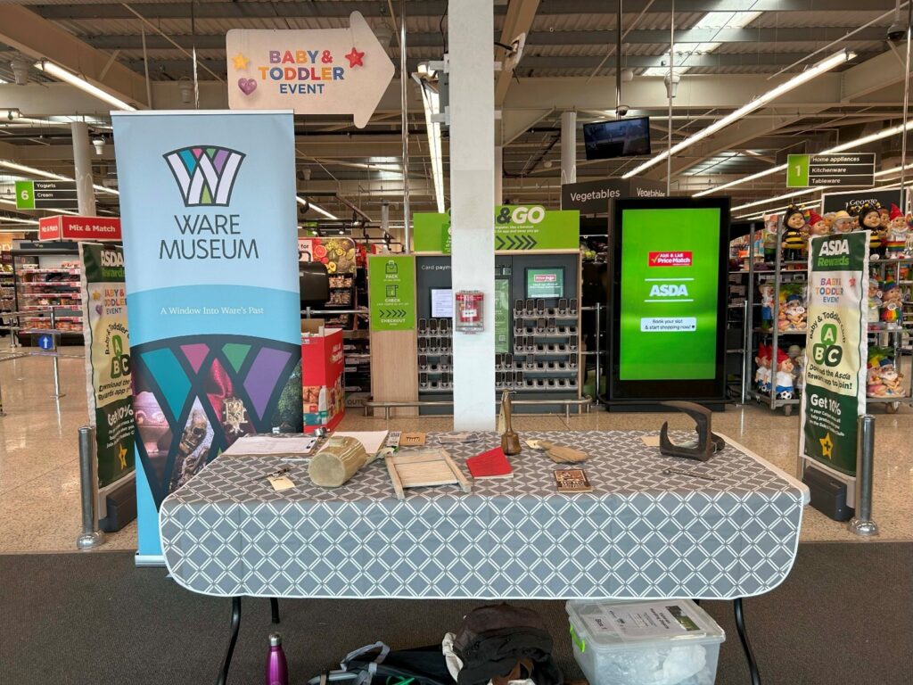 A table in front of the entrance to an Asda supermarket in Ware. The table has objects from the museum, a stone hot water bottle, a washboard, a cobblers last and a wooden herb crusher